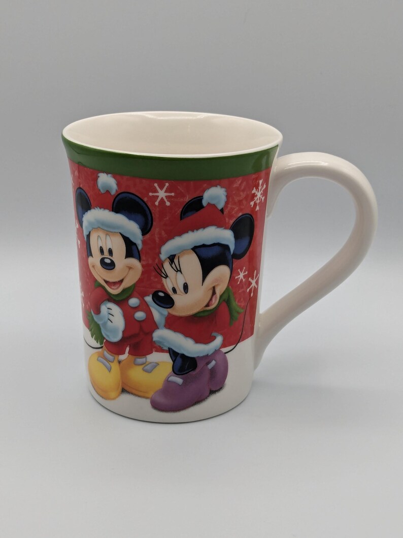 May include: A white ceramic mug featuring Mickey and Minnie Mouse in winter attire. The mug has a red background with snowflakes and a green rim. Mickey and Minnie are wearing red coats and hats.