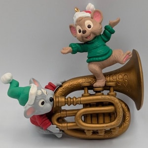 May include: A Christmas ornament featuring two cartoon mice. One mouse is standing on a gold-colored tuba, wearing a green sweater and Santa hat. The other mouse is wearing a red jacket and green hat. The year "1996" is engraved on the tuba.