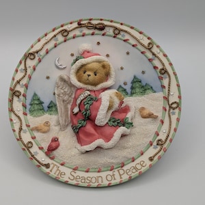 Könnte beinhalten: Ein dekorativer Weihnachtsteller mit einem Teddybär-Engel in rotem Gewand und Hut, der ein Lamm hält. Der Teller zeigt eine Winterlandschaft mit Vögeln, Bäumen und dem Text "The Season of Peace" am Rand.