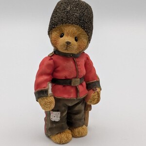 Puede incluir: Una figura de un oso de peluche marrón claro vestido como un guardia real británico. El oso lleva un gorro alto de piel de oso negro texturizado, una túnica roja con un cinturón negro y pantalones de color marrón oscuro con un pequeño parche blanco en la pierna derecha.