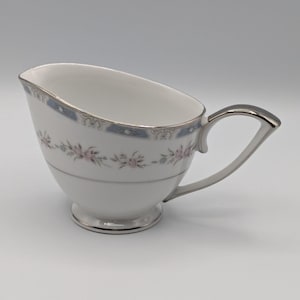 May include: White porcelain creamer with a silver handle and trim. The creamer features a delicate floral pattern in pink and gray, with a light blue border. The creamer has a small footed base.