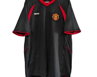 May include: Black short-sleeve jersey with red trim and accents. Features a V-neck collar and the MUFC logo on the chest. The Manchester United crest is on the right chest.