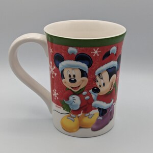 May include: A white ceramic mug featuring Mickey and Minnie Mouse in winter attire. The mug has a red background with white snowflakes and a green border. Mickey and Minnie are wearing red coats and hats.