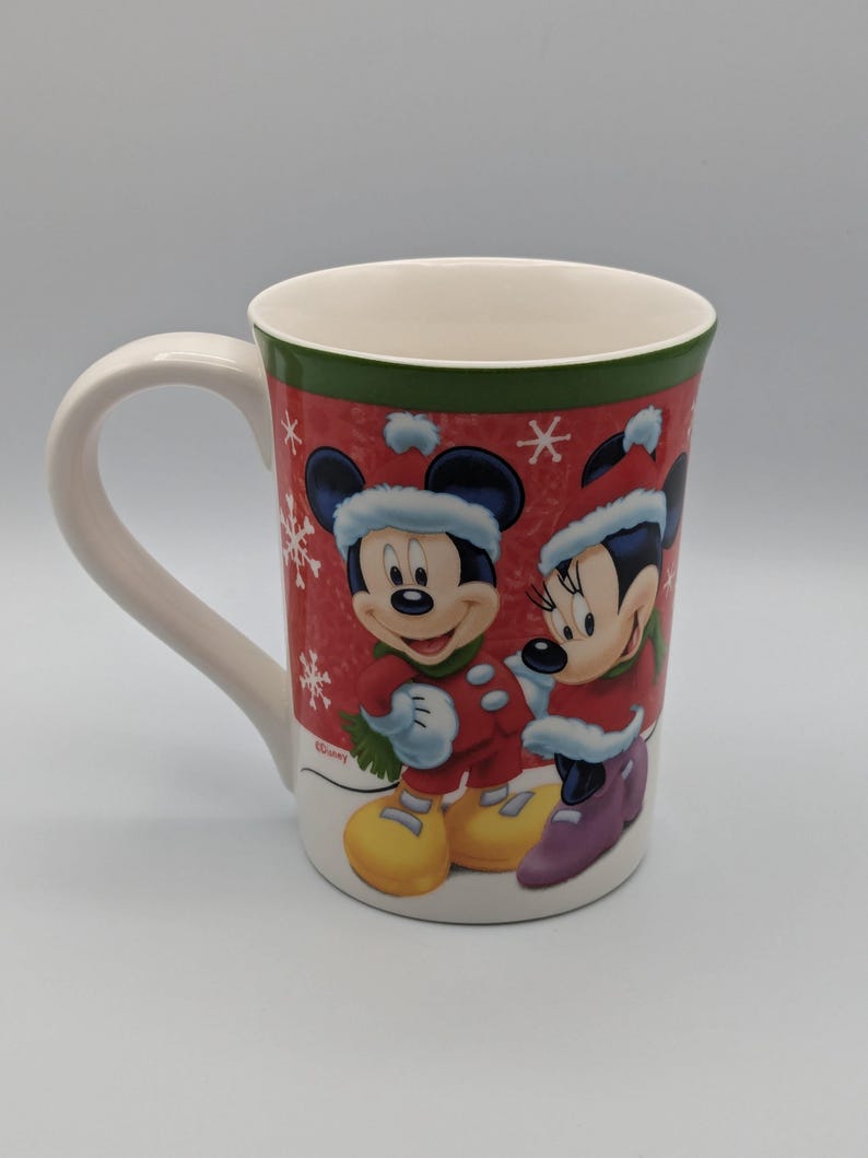May include: A white ceramic mug featuring Mickey and Minnie Mouse in winter attire. The mug has a red background with white snowflakes and a green border. Mickey and Minnie are wearing red coats and hats.