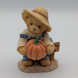 May include: A figurine of a teddy bear wearing a straw hat and denim overalls, holding a large orange pumpkin. The bear stands on a small, textured base, with a sign that reads "kins ck".
