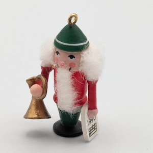 May include: A small wooden nutcracker ornament featuring a green hat, a red jacket, and green trousers. The figure has white faux fur hair and a beard, holds a gold-colored trumpet in one hand, and carries a white sign with the year 1990 printed on it.