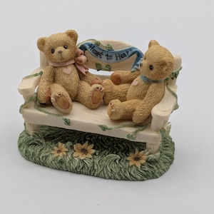 May include: A figurine of two teddy bears sitting on a white bench with a blue banner that reads "Heart to Heart." The bears are light brown with pink and blue ribbons. The base is green with yellow flowers.