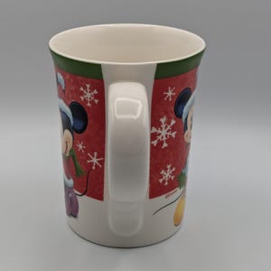May include: White ceramic mug with a red background featuring Mickey Mouse in a winter outfit, snowflakes, and a green rim. The mug has a white handle and is decorated with a winter theme.