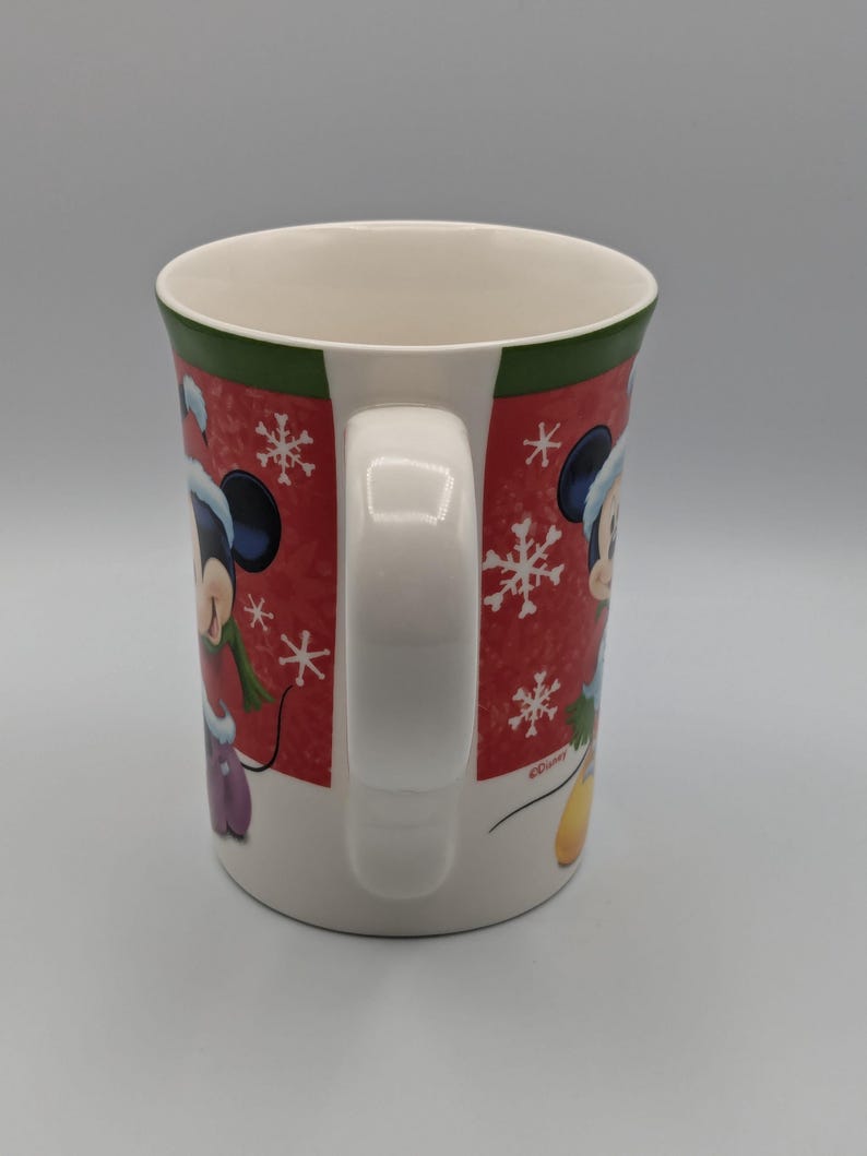 May include: White ceramic mug with a red background featuring Mickey Mouse in a winter outfit, snowflakes, and a green rim. The mug has a white handle and is decorated with a winter theme.