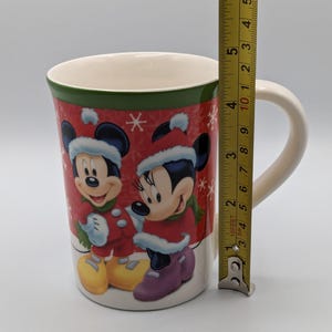 May include: A ceramic mug featuring Mickey and Minnie Mouse in winter attire. The mug has a red background with snowflakes and a green band at the top. The mug is approximately 10 cm tall.