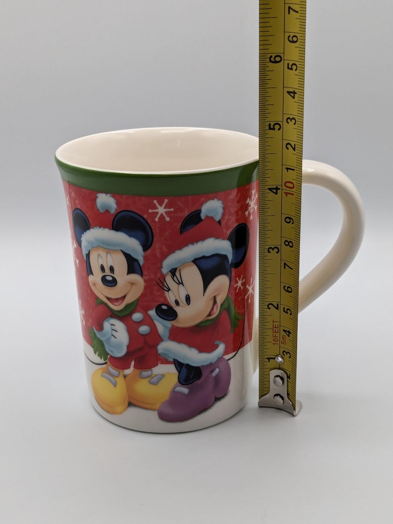 May include: A ceramic mug featuring Mickey and Minnie Mouse in winter attire. The mug has a red background with snowflakes and a green band at the top. The mug is approximately 10 cm tall.