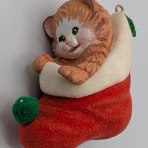 May include: A ceramic Christmas ornament featuring a ginger tabby cat nestled in a red stocking with a white cuff. The cat has green eyes and a smiling expression. A green pom-pom adorns the stocking's toe, and a gold loop is at the top for hanging.