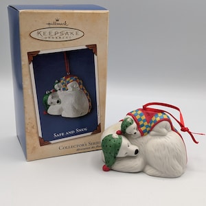 Könnte beinhalten: Ein Hallmark Keepsake-Ornament mit zwei Eisbären, die sich aneinander kuscheln. Einer trägt eine grüne Mütze, der andere eine rot-gelbe Decke. Auf der Box steht "Safe and Snug" und "Collector's Series".