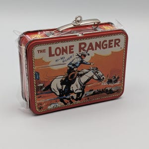 May include: A red metal lunchbox featuring a vintage illustration of The Lone Ranger riding a white horse, with the text "The Lone Ranger" and "Hi-Yo Silver!". The lunchbox has a silver handle and clasp.