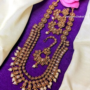 May include: A gold-tone necklace with multiple strands of pearls and clear stones. The necklace is layered with a matching maang tikka, earrings, and nose ring. The jewelry is displayed on a purple background.