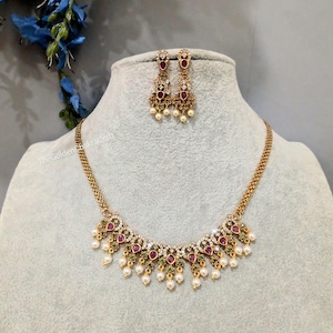 May include: A gold-toned necklace and matching earrings set. The necklace and earrings feature red and green gemstones, clear crystals, and small white pearls. The jewelry is displayed on a mannequin head against a gray background.