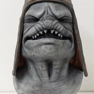 May include: A gray monster mask with a brown leather-like cap. The mask has a wrinkled face, sharp teeth, and a menacing expression. The mask is mounted on a black stand, suitable for Halloween or cosplay.