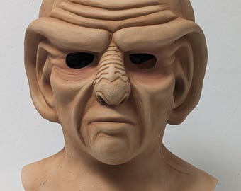 May include: A tan-colored mask with a detailed, wrinkled face, prominent brow ridges, and large ears. The mask has dark eye sockets and a textured surface, mounted on a black stand. The mask is likely a costume or prop.
