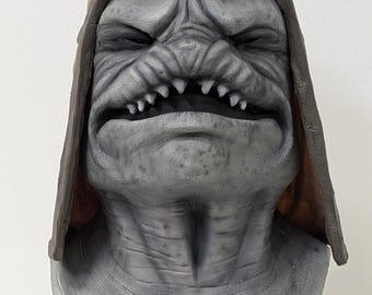 May include: A gray monster mask with a brown leather-like cap. The mask has a wrinkled face, sharp teeth, and a menacing expression. The mask is mounted on a black stand, suitable for Halloween or cosplay.