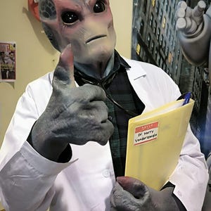 May include: An alien doctor in a white lab coat gives a thumbs up. The alien has gray skin, large black eyes, and a red area around the eyes. The doctor is holding a yellow folder with a name tag that reads "Dr. Harry Vanderspeigle."