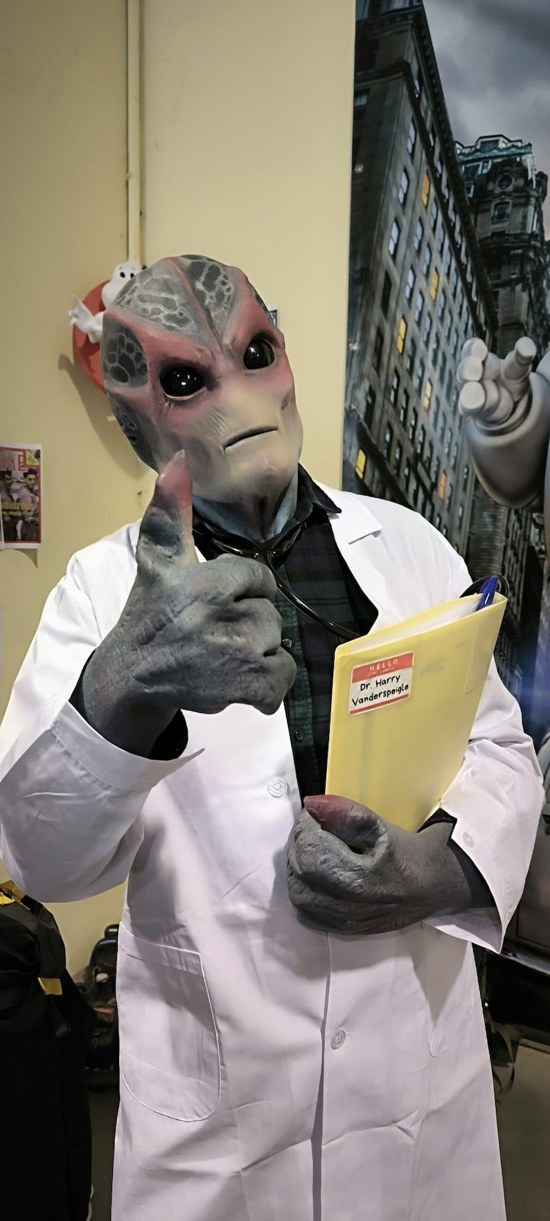 May include: An alien doctor in a white lab coat gives a thumbs up. The alien has gray skin, large black eyes, and a red area around the eyes. The doctor is holding a yellow folder with a name tag that reads "Dr. Harry Vanderspeigle."