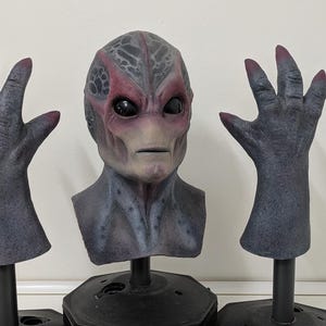 May include: A detailed alien mask and two matching hand props. The mask is gray and red with large black eyes. The hands are gray with red fingernails. All items are mounted on black stands, perfect for cosplay or Halloween.