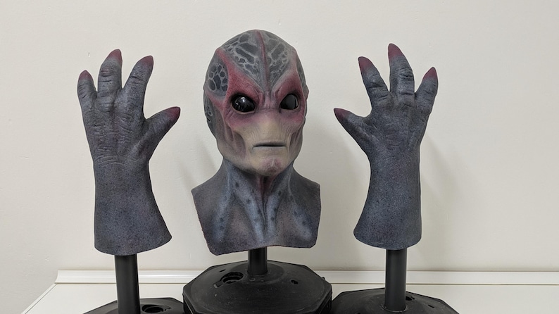 May include: A detailed alien mask and two matching hand props. The mask is gray and red with large black eyes. The hands are gray with red fingernails. All items are mounted on black stands, perfect for cosplay or Halloween.