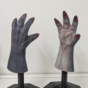 May include: Two gray, articulated monster hands with dark red fingernails are displayed on black stands. The hands are posed with fingers spread, showcasing a detailed, textured skin effect, perfect for Halloween.