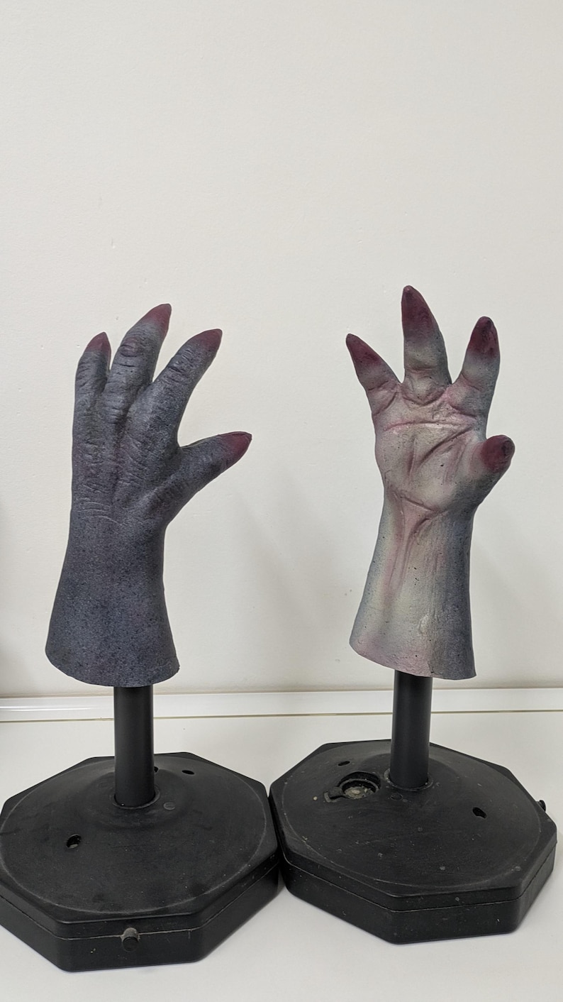 May include: Two gray, articulated monster hands with dark red fingernails are displayed on black stands. The hands are posed with fingers spread, showcasing a detailed, textured skin effect, perfect for Halloween.
