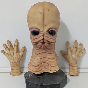 Puede incluir: Una máscara alienígena de color beige con grandes ojos negros y un cuello acanalado. Dos manos beige a juego con venas detalladas se colocan a cada lado. La máscara está montada sobre una base hexagonal negra.