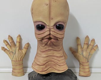 May include: A tan alien mask with large black eyes and a ridged neck. Two matching tan hands with detailed veins are positioned on either side. The mask is mounted on a black hexagonal base.