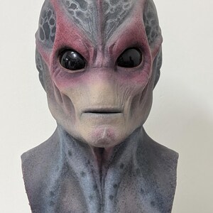 May include: A detailed alien mask with a textured, gray and red skin tone. The mask features large, black eyes and a neutral expression. The mask is mounted on a black stand, suggesting it's a display piece or costume accessory.