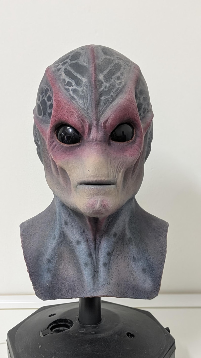 May include: A detailed alien mask with a textured, gray and red skin tone. The mask features large, black eyes and a neutral expression. The mask is mounted on a black stand, suggesting it's a display piece or costume accessory.