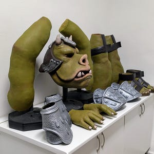 May include: A collection of green and silver costume pieces, including a monster mask with horns, arms, legs, hands, and armor. The mask is brown and green with white teeth. The armor is silver. The costume pieces are displayed on a white surface.