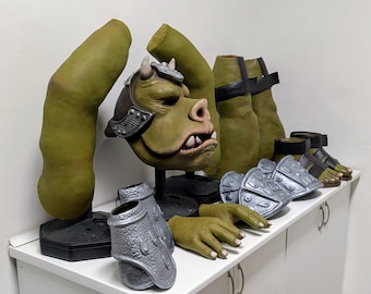 May include: A collection of green and silver costume pieces, including a monster mask with horns, arms, legs, hands, and armor. The mask is brown and green with white teeth. The armor is silver. The costume pieces are displayed on a white surface.