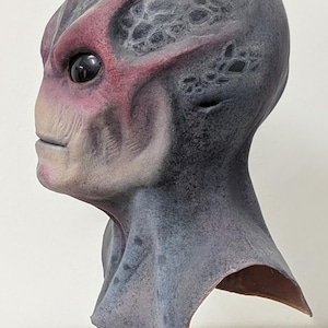 May include: A detailed alien mask with a gray and purple color scheme. The mask features a prominent forehead, large black eyes, and a textured surface. The alien mask is mounted on a black stand.