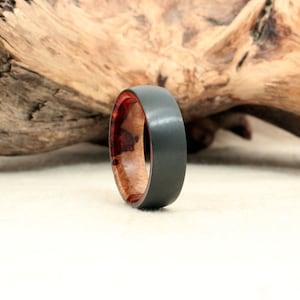 May include: A black and gray tungsten carbide ring with a red and brown wood inlay. The ring is shown on a wooden background.