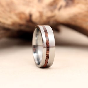 Puede incluir: Un anillo de titanio plateado con una incrustación de madera marrón. El anillo tiene un diseño simple con una sola banda de madera.