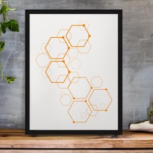 Könnte beinhalten: Gerahmter Kunstdruck mit einem abstrakten Design aus orangefarbenen Sechsecken auf weißem Hintergrund. Das Kunstwerk befindet sich in einem schwarzen Rahmen und steht auf einer Holzoberfläche. Eine Rankpflanze und eine kleine Vase mit einer Pflanze sind ebenfalls zu sehen.