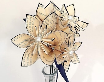 May include: Three paper flowers made from book pages with pearl accents, arranged in a clear glass vase with a navy blue ribbon.