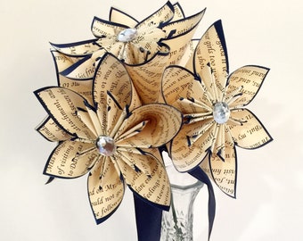 May include: A bouquet of paper flowers made from old book pages, with navy blue ribbon and rhinestones, in a clear glass vase.