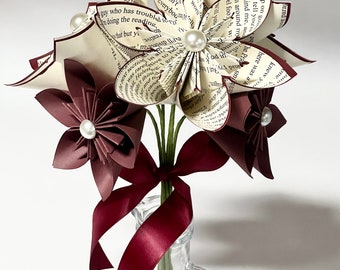 May include: A bouquet of paper flowers made from book pages, with burgundy paper flowers and white pearl accents. The bouquet is tied with a burgundy ribbon and placed in a clear glass vase.