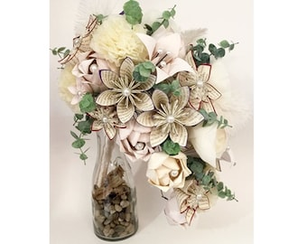 May include: A cascading bouquet of paper flowers in shades of white and cream. The bouquet is made with book pages, fabric, and greenery. It is displayed in a clear glass vase with small stones.