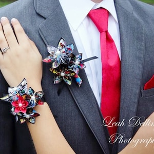 May include: A gray suit jacket with a red tie and a red pocket square. The jacket has a comic book themed boutonniere and a matching wrist corsage.