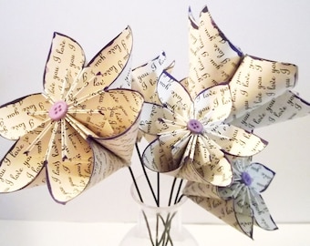 May include: A bouquet of five paper flowers made from old book pages. The flowers are white and cream with purple buttons in the center. The flowers are arranged in a clear glass vase.
