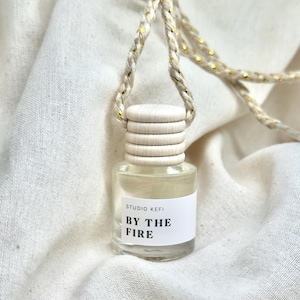 May include: A small, clear glass bottle with a wooden cap and a woven cord for hanging. The label reads "BY THE FIRE" in black text. The cord has gold accents. The bottle is likely an air freshener or essential oil diffuser.