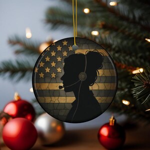May include: A round Christmas ornament featuring a black silhouette of a person wearing a headset, set against a distressed American flag design in gold and black. The ornament hangs from a gold string, perfect for holiday decor.