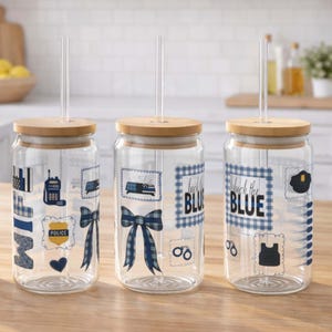 May include: Three clear glass tumblers with bamboo lids and straws. Each tumbler features blue and white graphics, including a police badge, bow tie, and the text "Back the Blue". The tumblers are on a wooden surface.