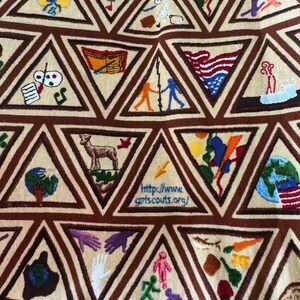 Puede incluir: Primer plano de una tela beige con tri&aacute;ngulos marrones bordados. Cada tri&aacute;ngulo contiene una imagen bordada colorida diferente, que incluye una bandera estadounidense, un ciervo y un globo. La direcci&oacute;n del sitio web http://www.girlscouts.org/ es visible.