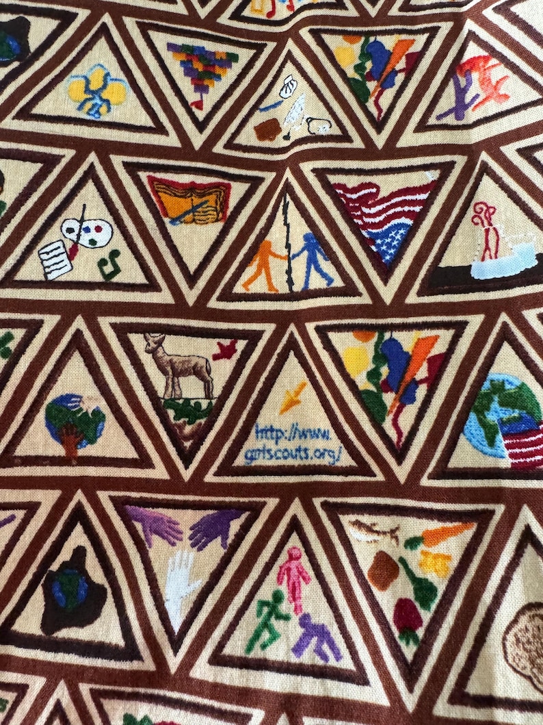 Puede incluir: Primer plano de una tela beige con tri&aacute;ngulos marrones bordados. Cada tri&aacute;ngulo contiene una imagen bordada colorida diferente, que incluye una bandera estadounidense, un ciervo y un globo. La direcci&oacute;n del sitio web http://www.girlscouts.org/ es visible.